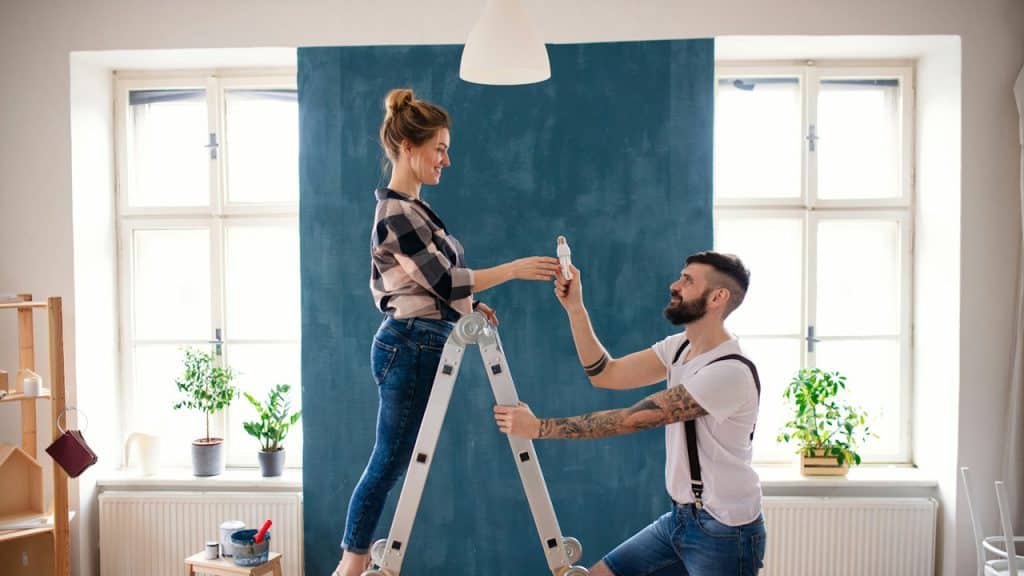 A woman standing on a ladder and a man handing her a small item.