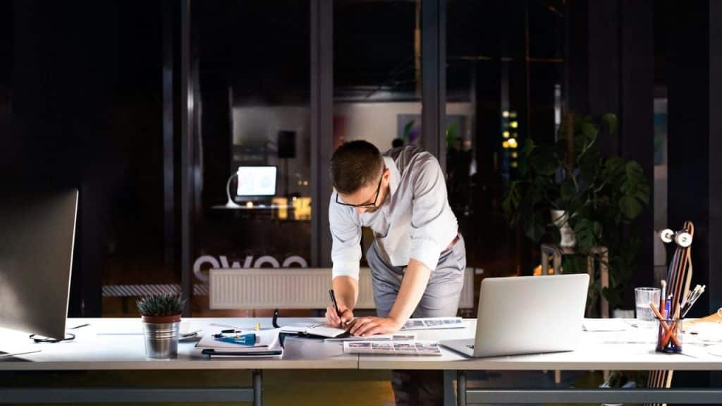 Man writing in a notebook, bending over a desk in a dark office at night.