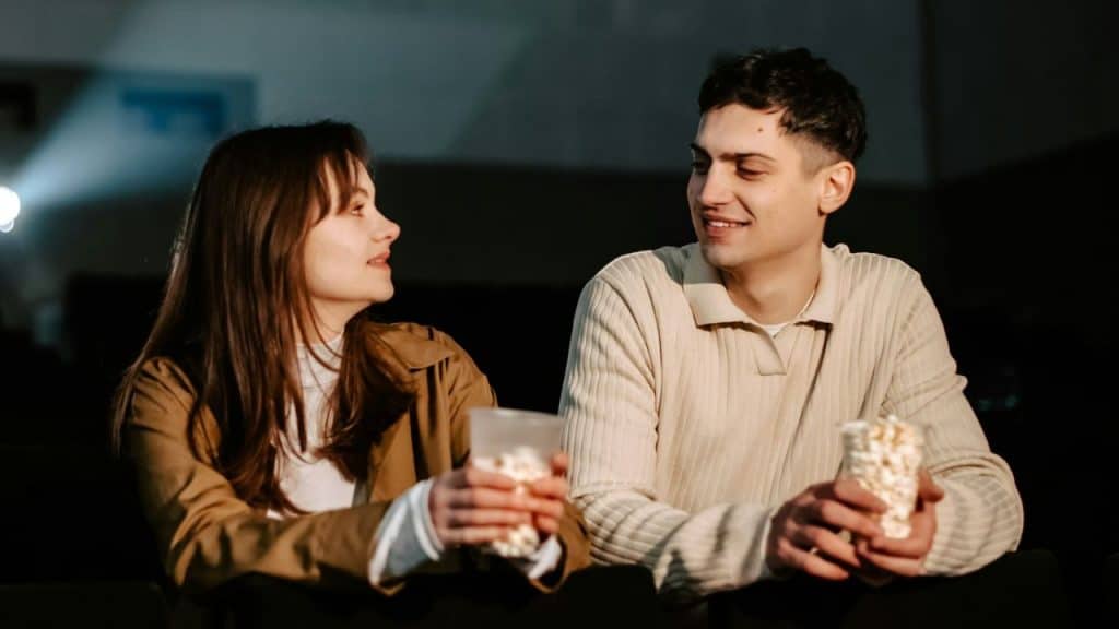 A happy man and woman talk while holding popcorn in a dimly lit movie theater.