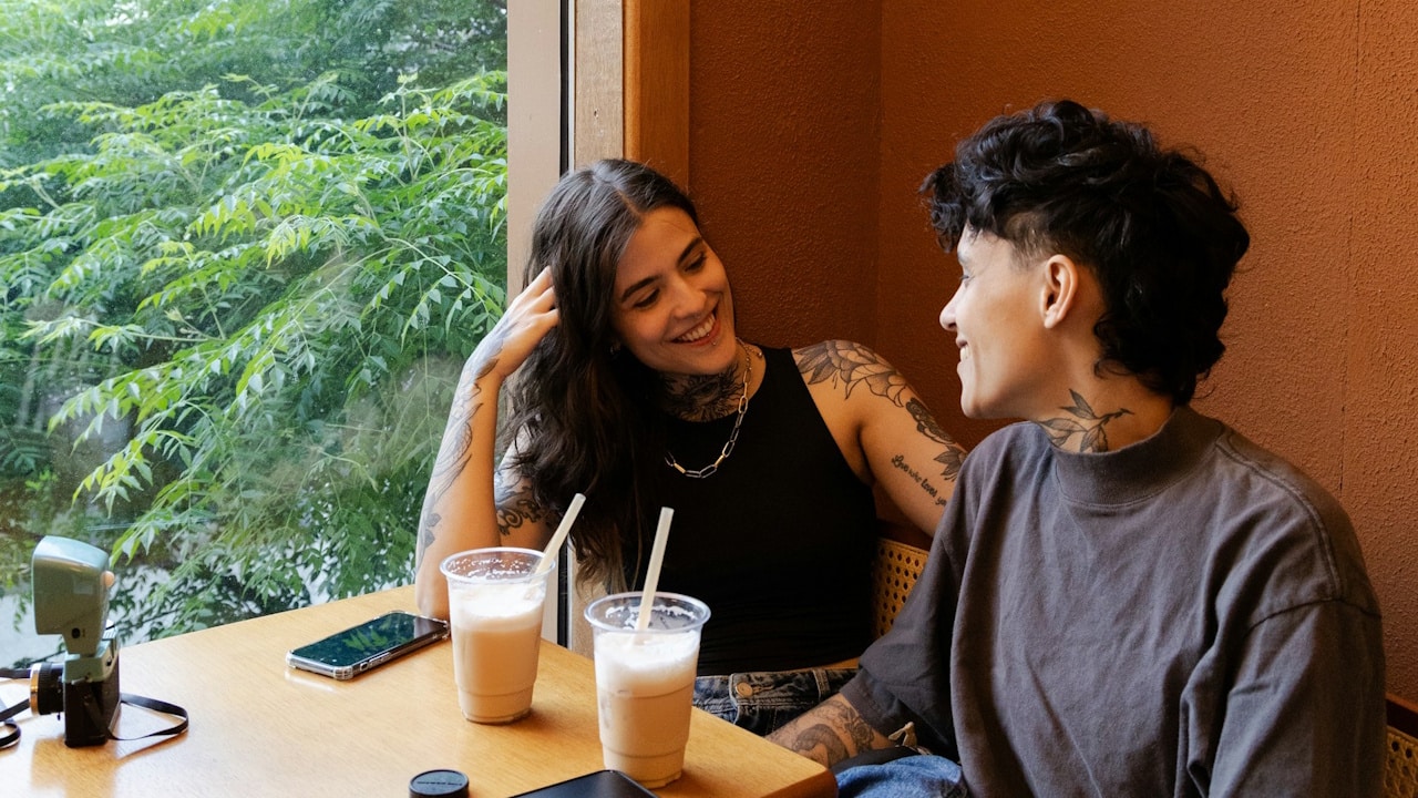 Two smiling, tattooed individuals sit at a table by a large window overlooking greenery, sharing iced drinks.