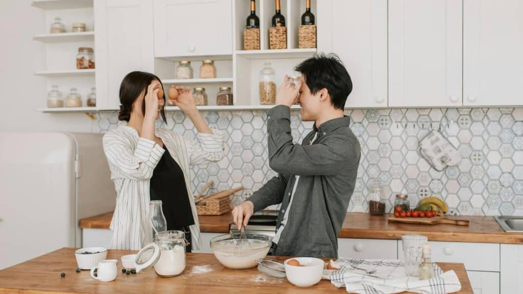 A couple playfully holding eggs in front of their faces while cooking together in a kitchen.