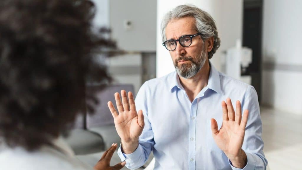 Mature man with a beard and glasses holding both hands up in a "stop" gesture while talking to a blurred person.