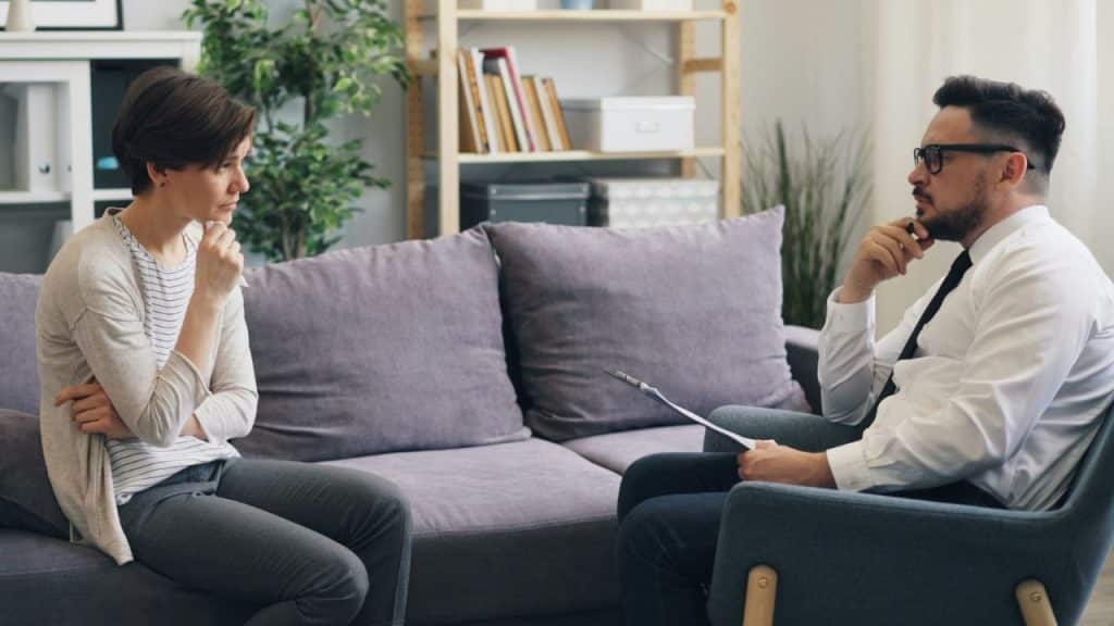 A thoughtful woman sits on a couch facing a mant in a white shirt.
