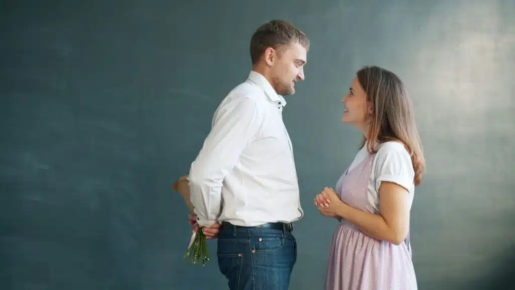 Man holding flowers behind his back while smiling at a woman in a light dress.