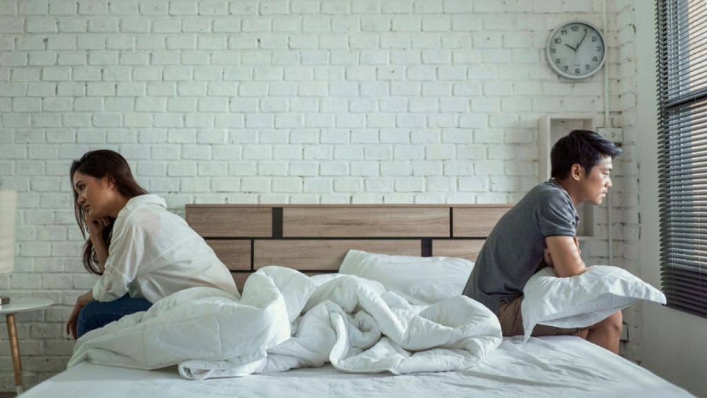 A man and a woman sitting on opposite sides of a bed, looking away from each other.