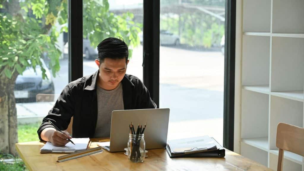 Young man working intently on a laptop and taking notes at a wooden desk near a window.