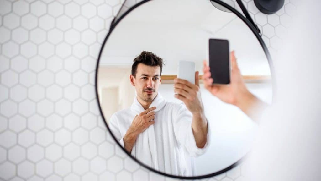 Man in a white bathrobe taking a selfie in a round mirror in a bathroom.