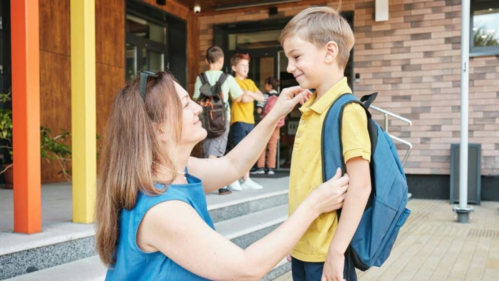 Woman kneeling and adjusting the collar of a boy with a blue backpack outside a school.