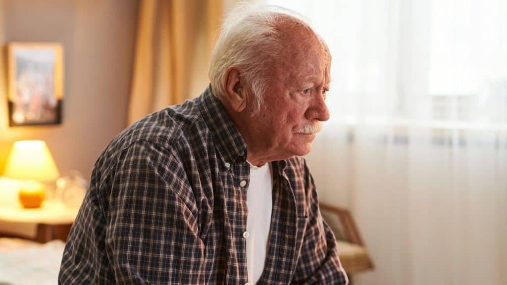 Older man in a plaid shirt looking downcast while sitting indoors.