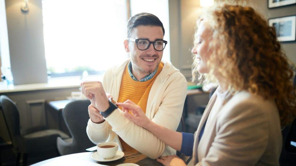 Man and woman sitting at a table smiling, looking at a smartwatch on the man's wrist.