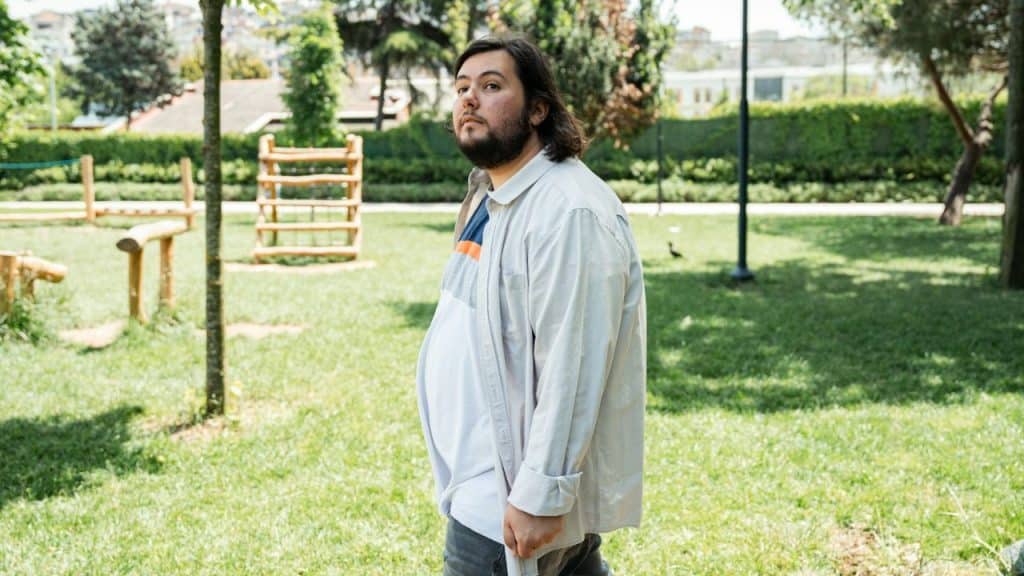 Man with a beard and long hair wearing an oversized shirt in a sunny park.
