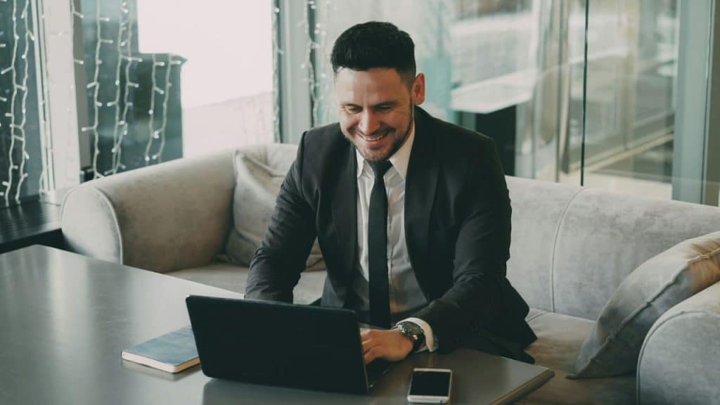 A smiling businessman working on a laptop while seated on a sofa indoors.