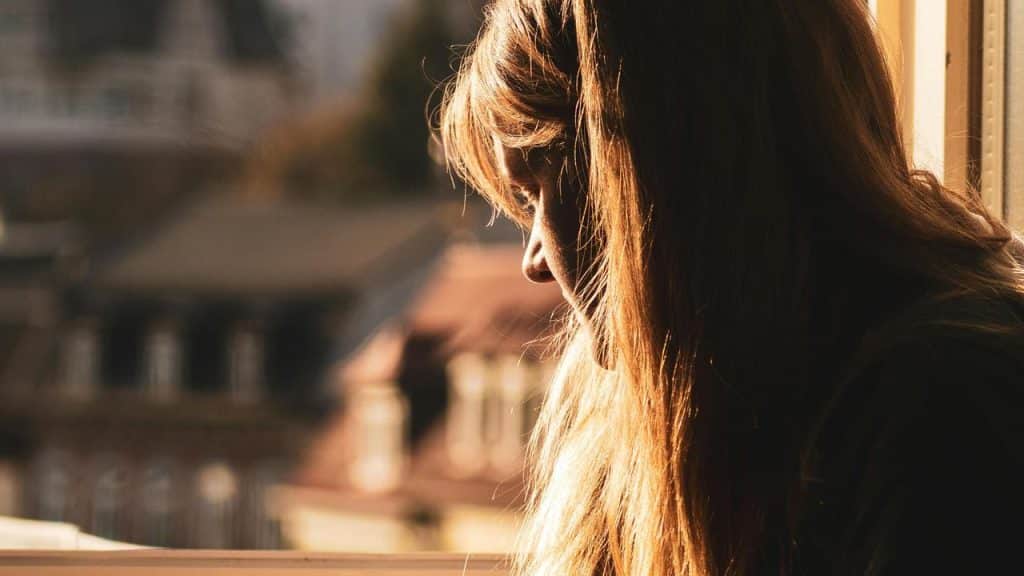 A woman sitting near a sunlit window, looking thoughtful.