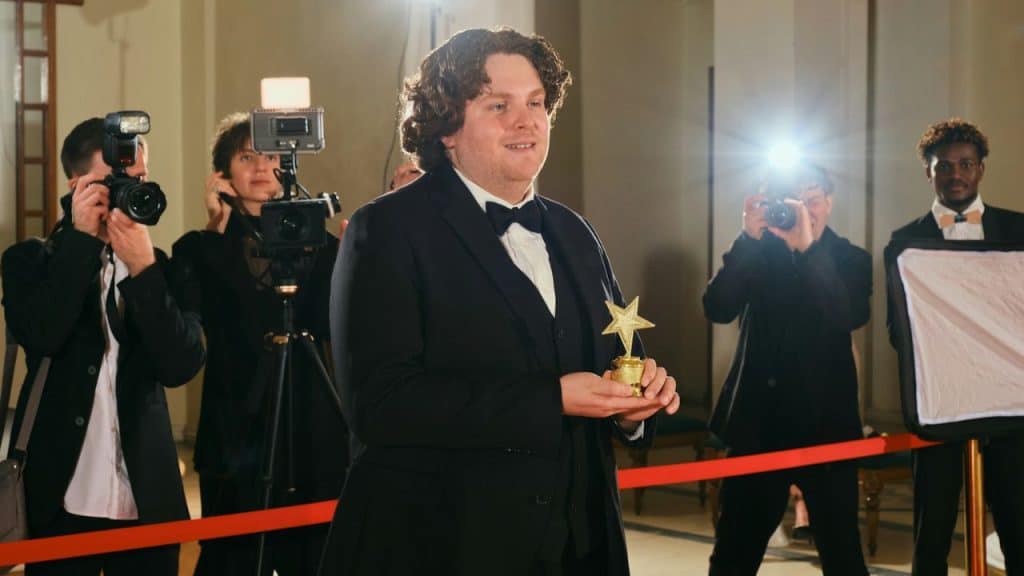 Man in a tuxedo holding a star trophy while being photographed by paparazzi.