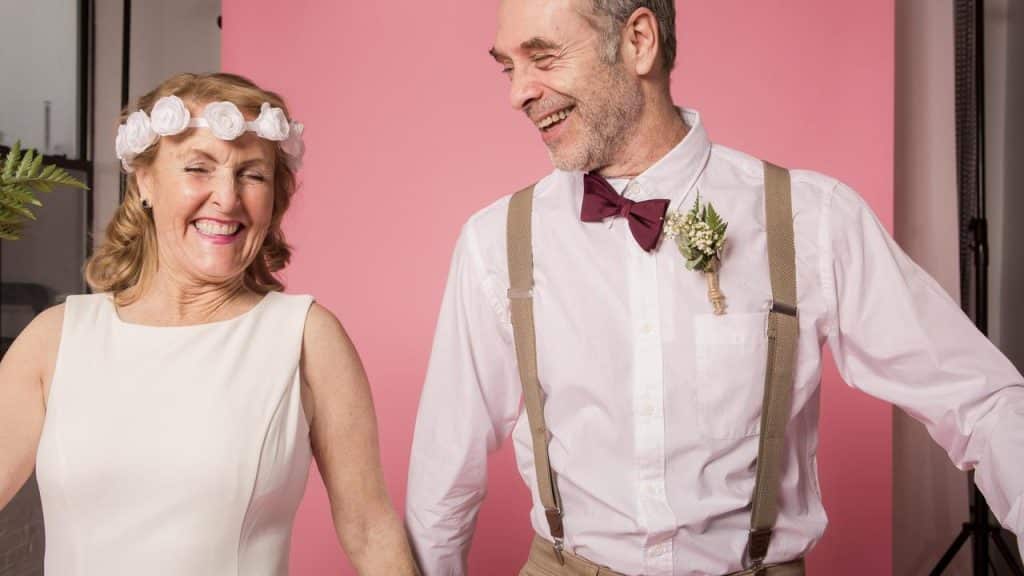 An older couple dressed for a wedding, smiling and holding hands against a pink background.