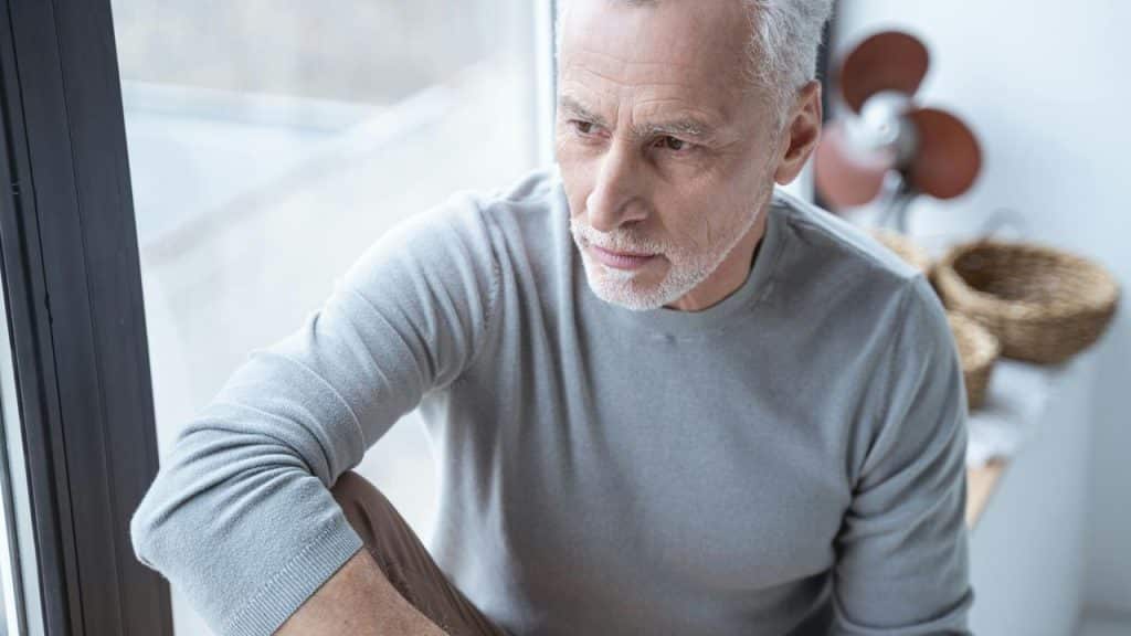 An older man sitting by a window, looking thoughtful.