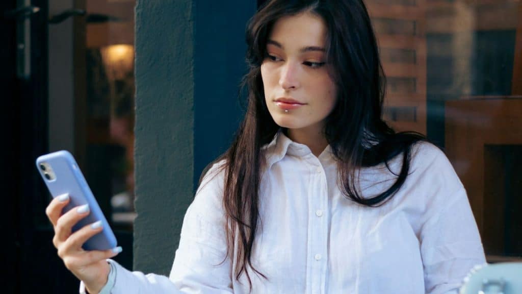 Woman with dark hair in a white shirt looking at a light blue smartphone outdoors.