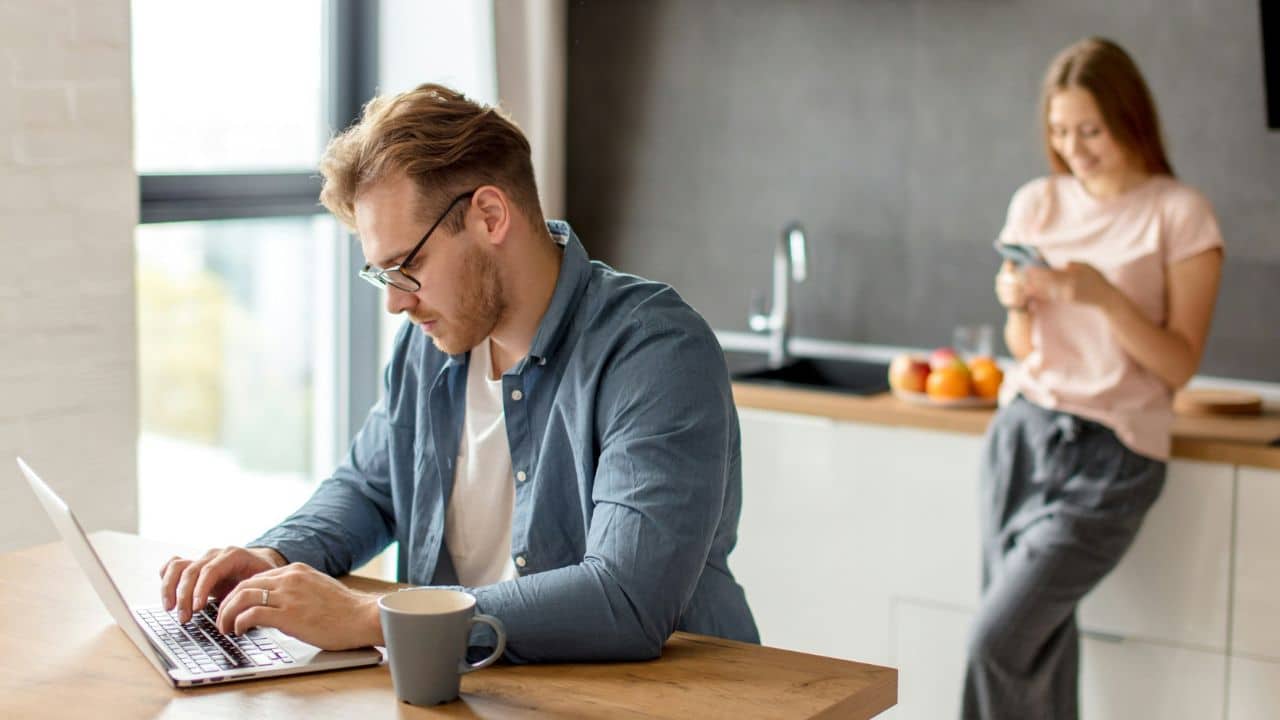 Man focused on a laptop at a kitchen counter while a woman uses her phone behind him.