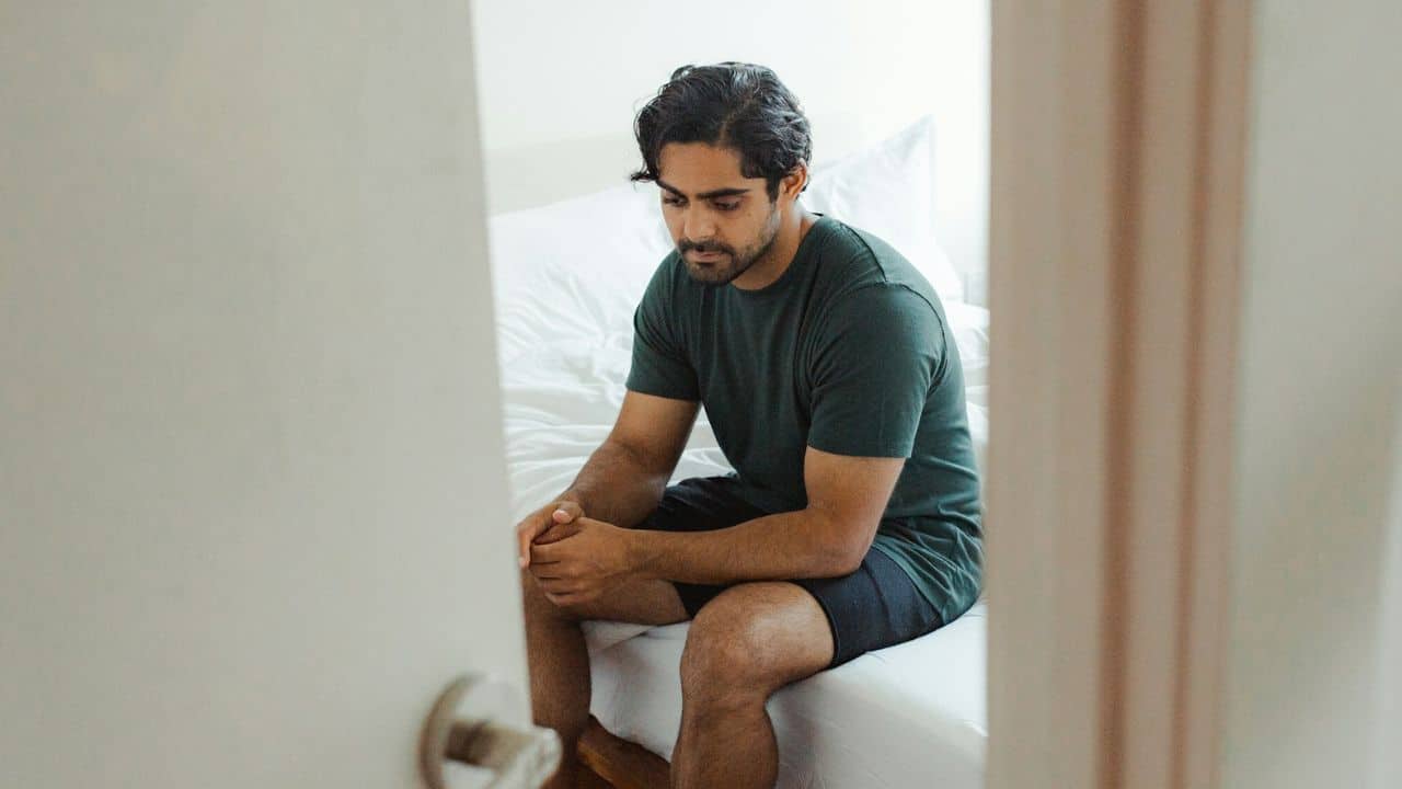 Man sitting on the edge of a bed, looking down, framed by a doorway.