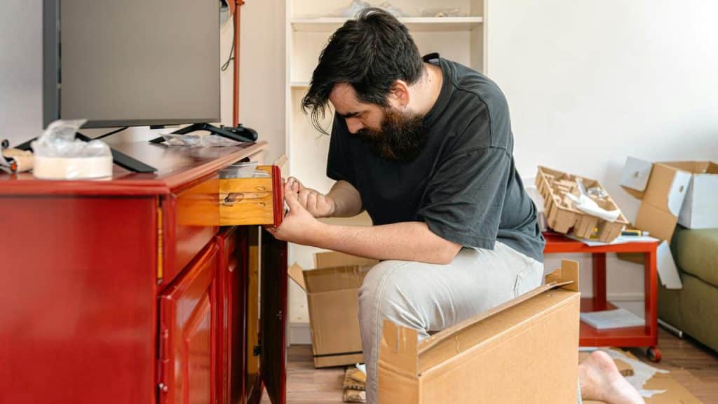 A man assembling a red wooden cabinet while sitting on the floor in a cluttered room.