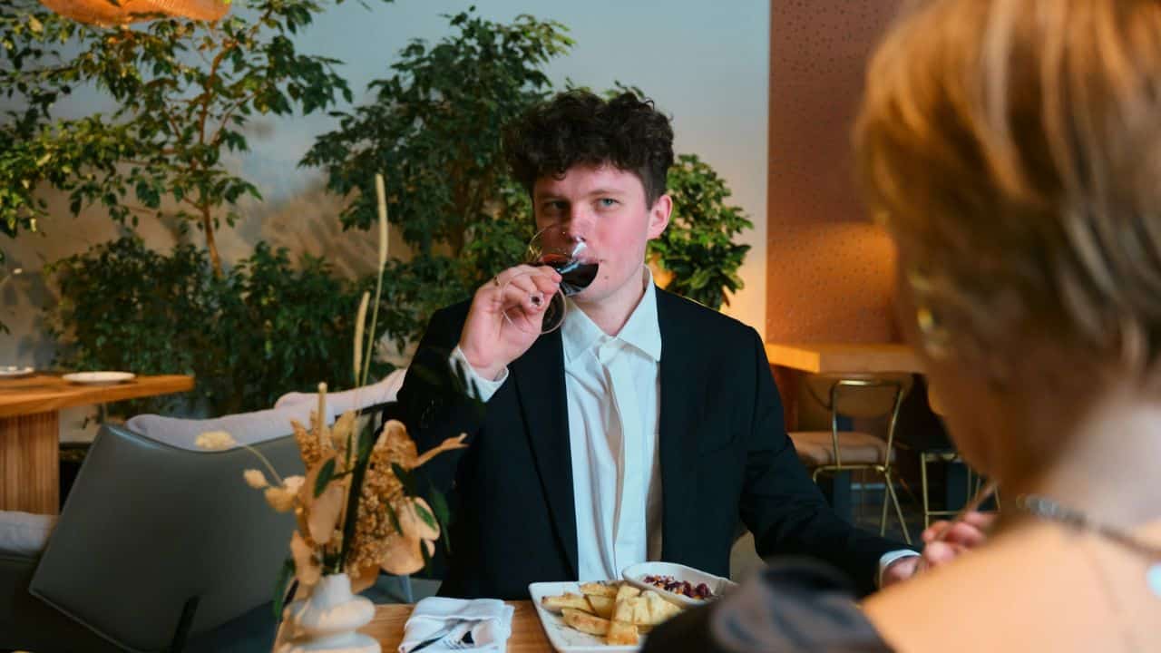 Young man in a black suit sips red wine at a restaurant table with a date.