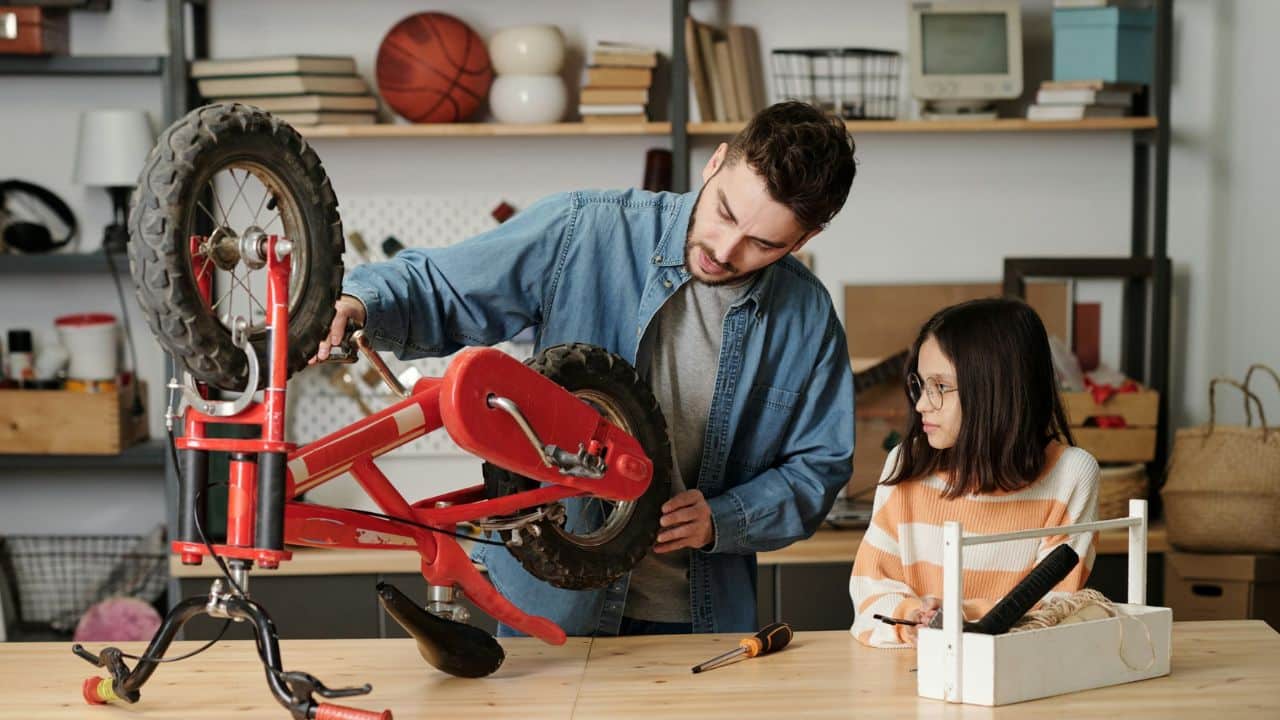 Man repairing a small red bicycle while a girl watches him from a table.