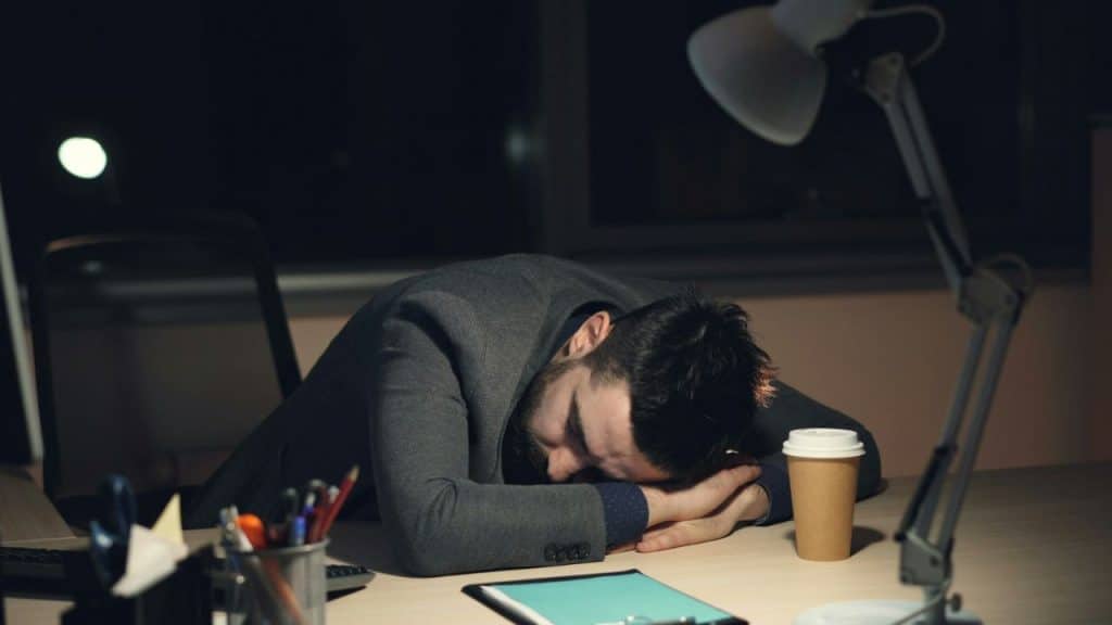 Tired man asleep face-down on a desk under a lamp, with a coffee cup.
