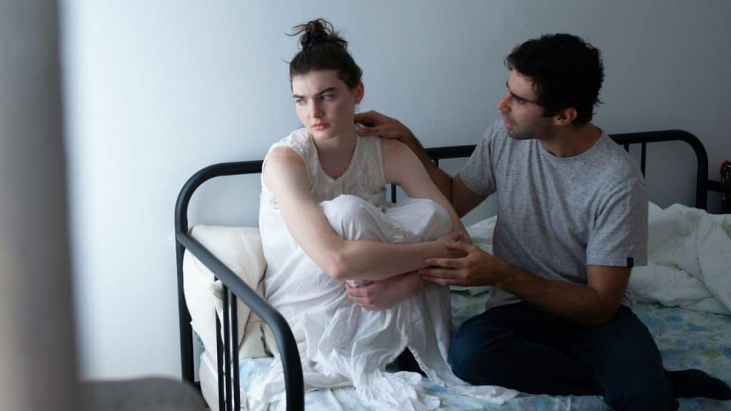 A sad woman sitting with knees up on a bed while a man comforts her.