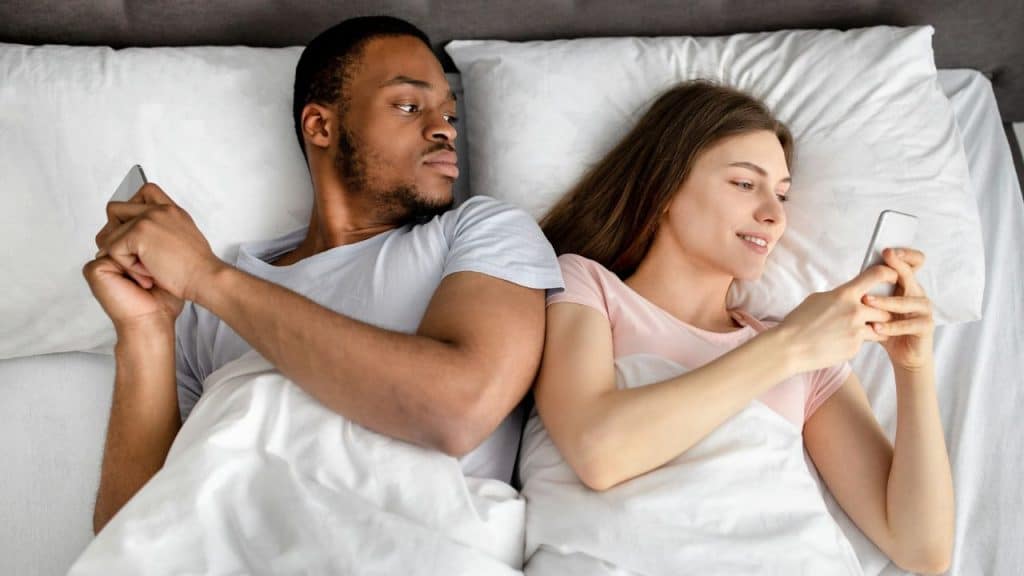 A man and woman in bed are on their phones; the man looks at the woman's screen.