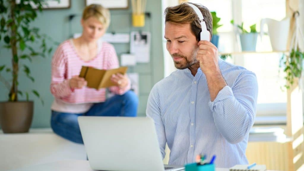 Man with headphones using a laptop, and a woman reading in the background.