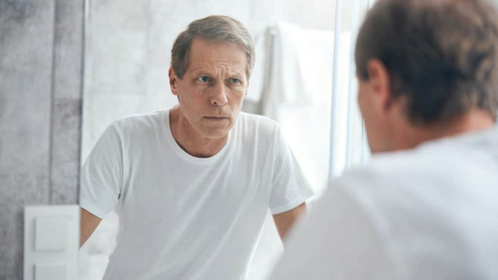 Middle-aged man in a white t-shirt looking intensely at his reflection in a bathroom mirror.