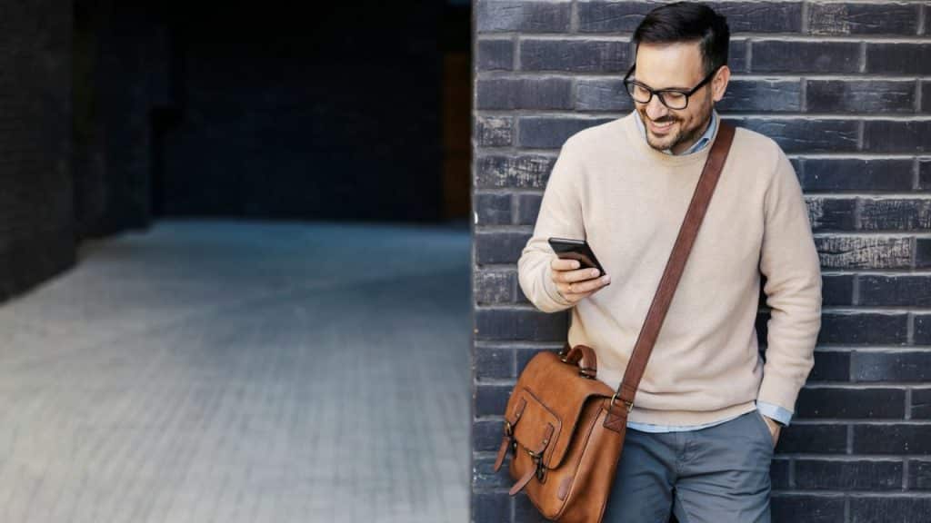 A smiling man in glasses, wearing a sweater and shoulder bag, looks at his cell phone.