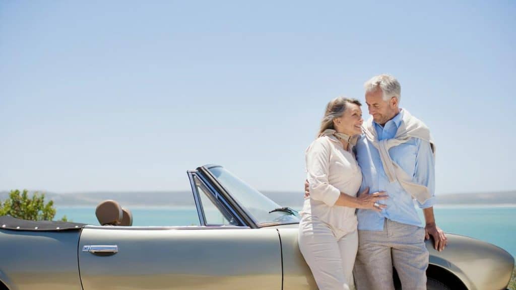 An older couple standing beside a convertible car near the ocean.