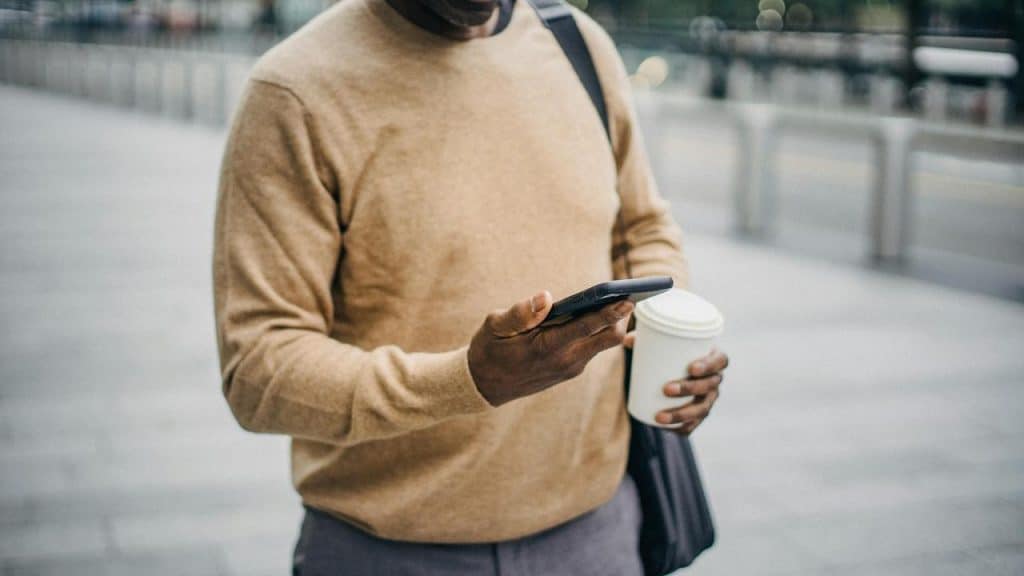 A man holding a coffee cup and looking at his phone while standing outdoors.
