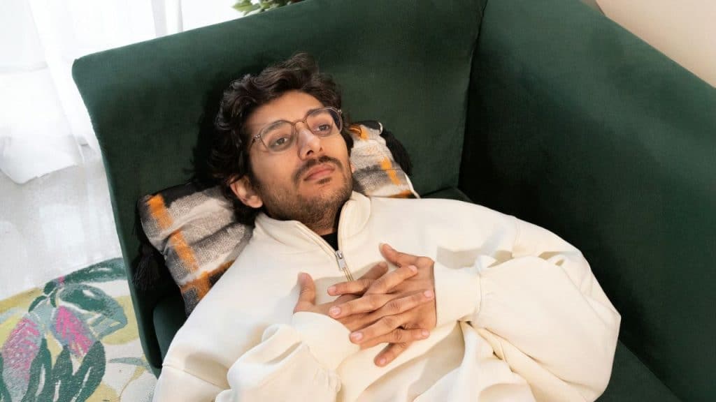Young man in a white sweatshirt and glasses lying on a dark green couch with his hands on his chest.