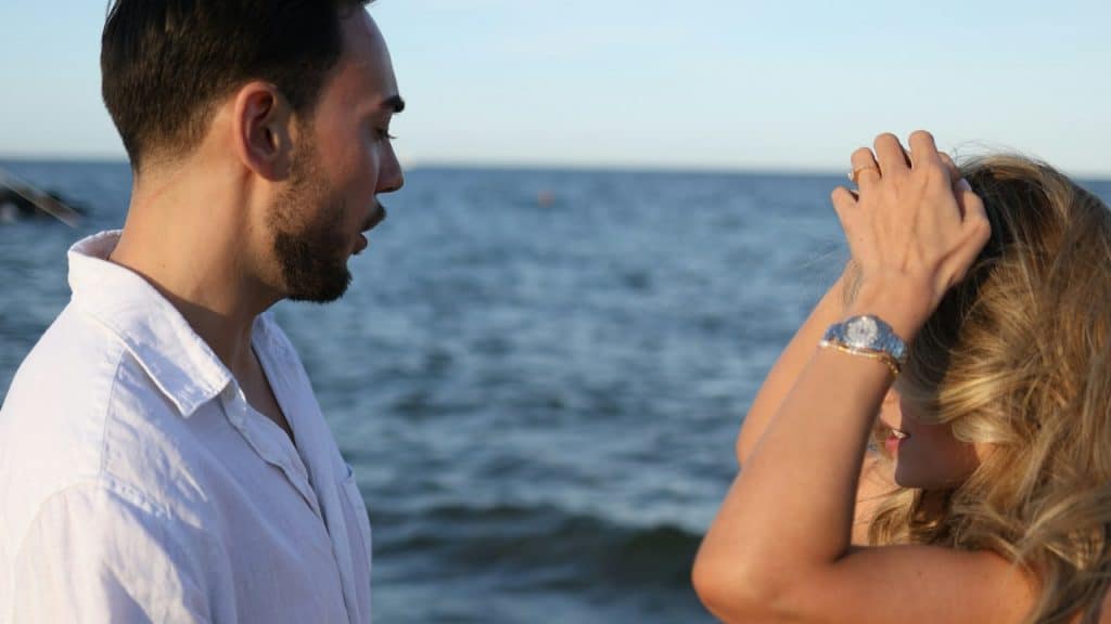A lightly bearded man and woman standing by the sea while the woman looks upset.
