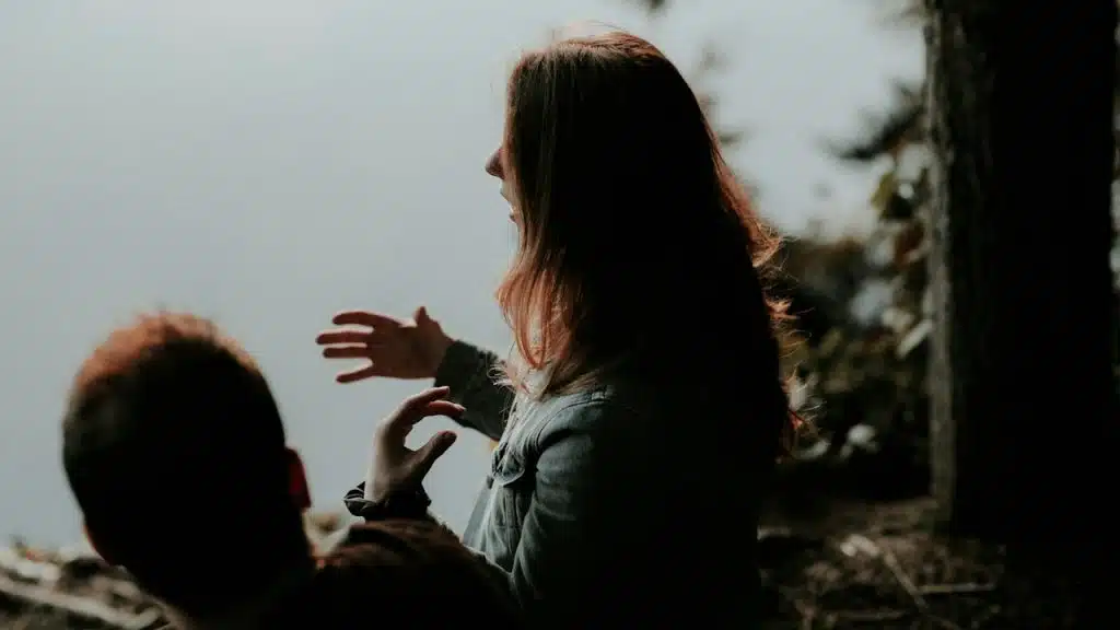 Woman gesturing with her hands while talking to a man outdoors by a foggy lake.