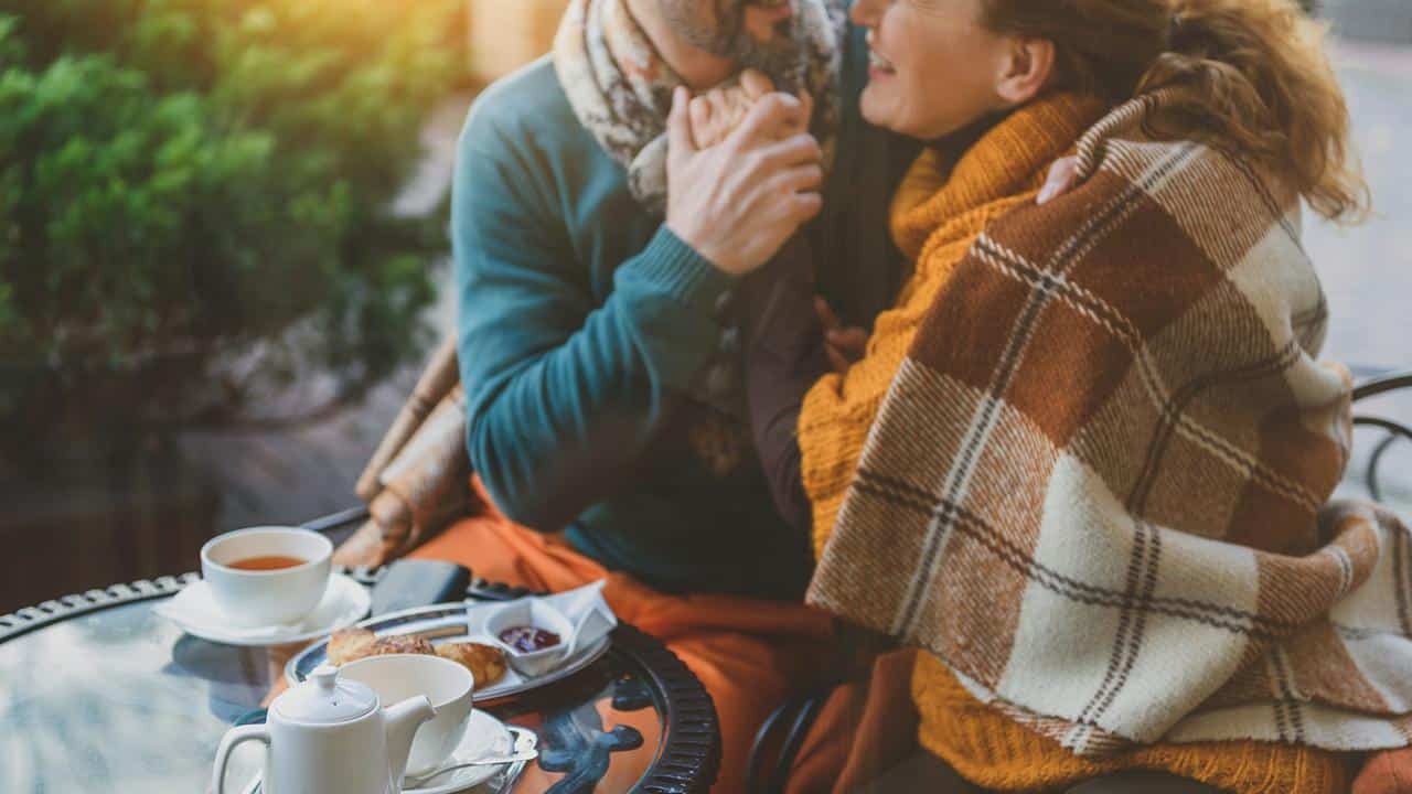 A couple sitting close together under a blanket, holding hands at an outdoor café.