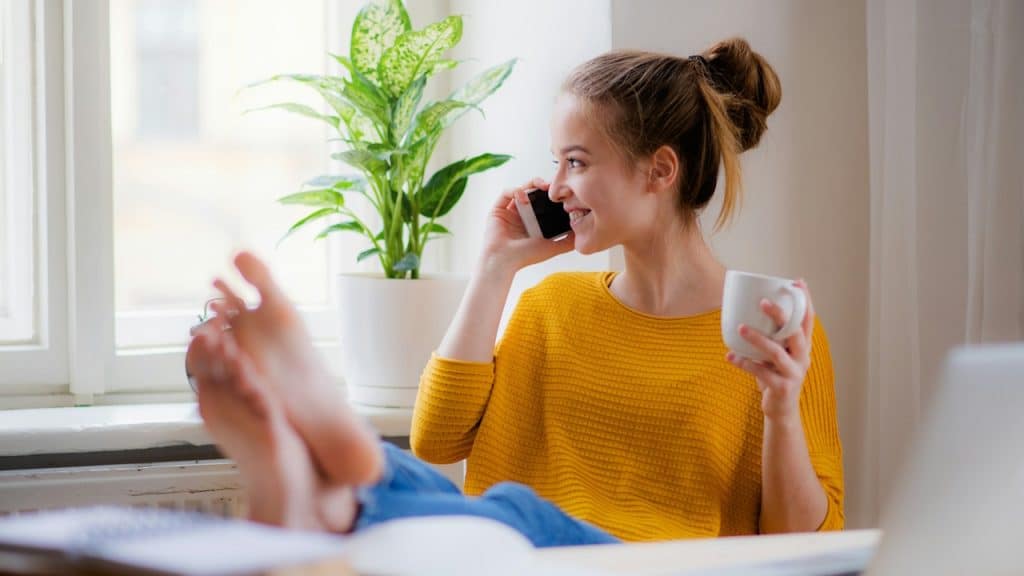 Smiling woman talking on a mobile phone, holding a mug, and relaxing with her feet up.