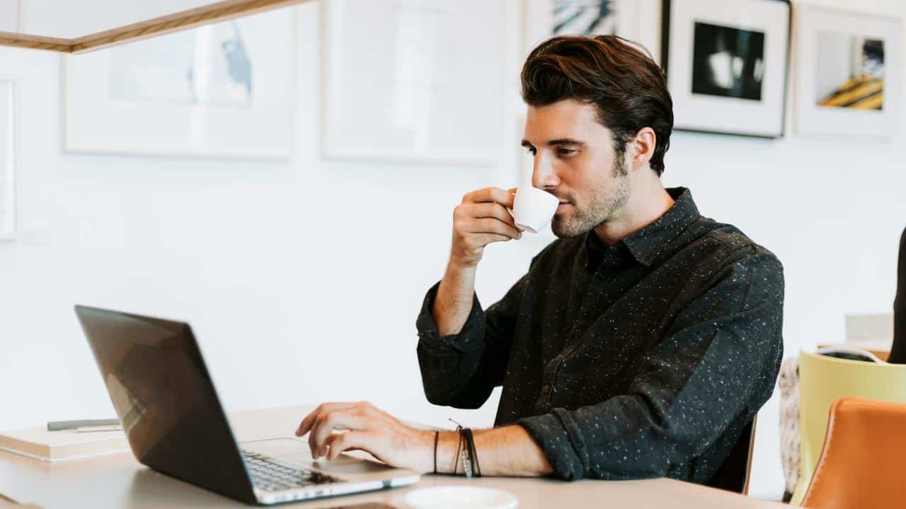 A man drinking coffee while working on a laptop at a desk.