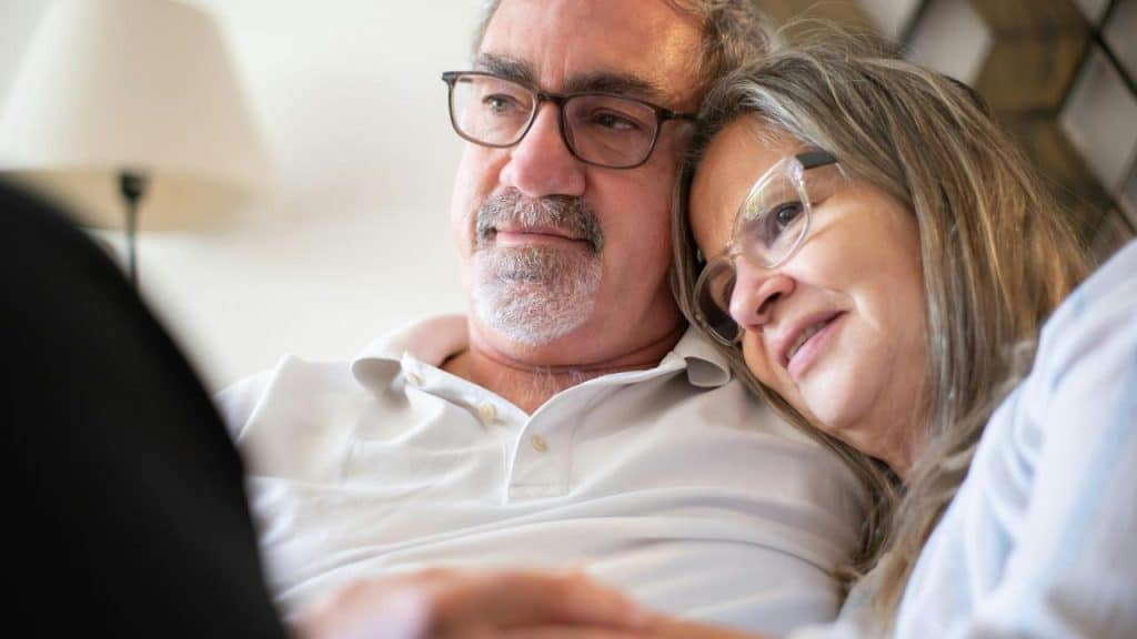 An older couple wearing glasses sitting close together and smiling warmly.