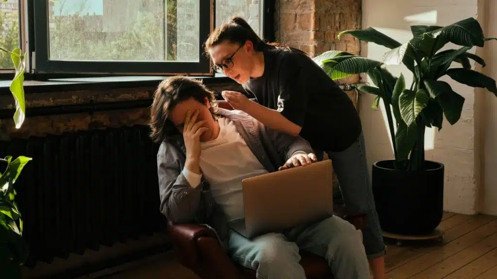 A distressed man covers his face at a laptop while a woman leans in and talks to him.