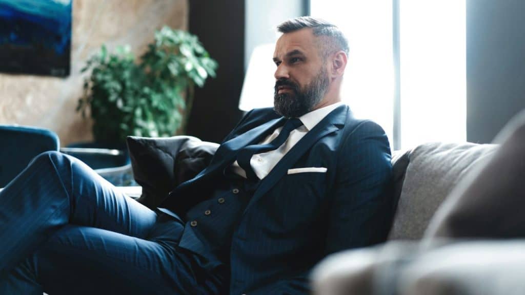 A well-dressed bearded man in a pinstripe suit reclines on a gray couch.