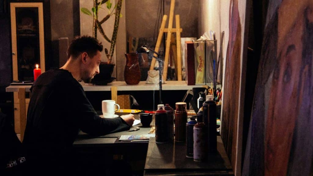 A man sits at a dimly lit desk cluttered with art supplies and a coffee mug.