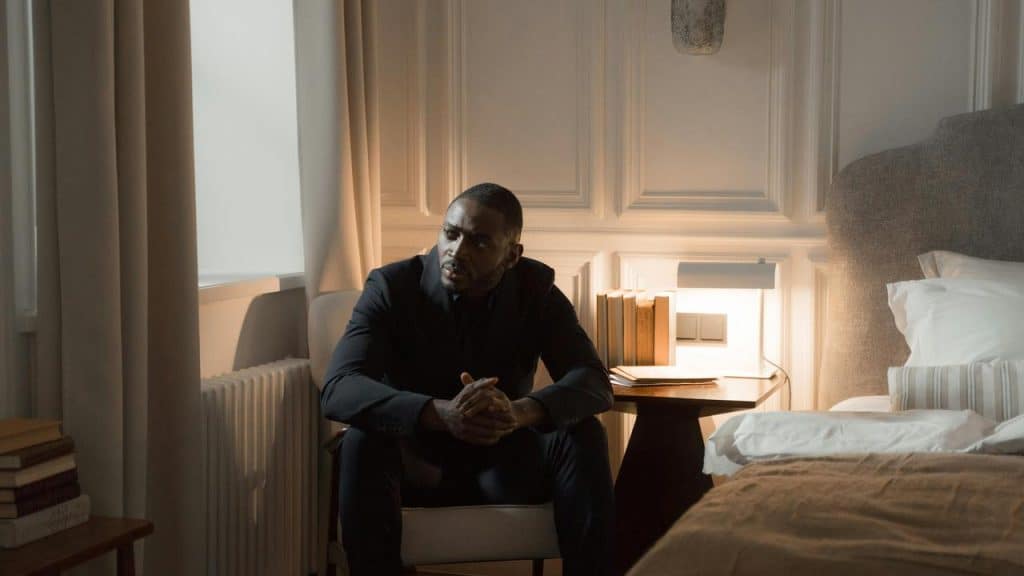 A man in a dark suit sits thoughtfully on a chair beside a bed in a softly lit bedroom.