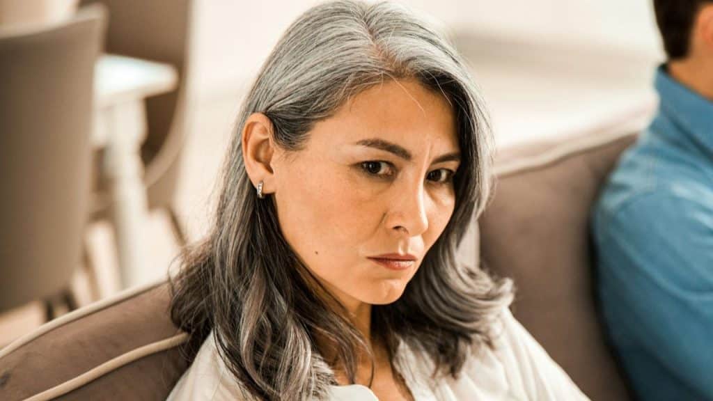 A woman with long gray hair and a serious expression sits on a couch indoors.
