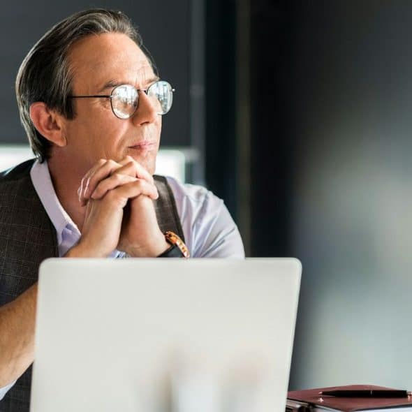 Older man in glasses and a vest, sitting at a desk with a laptop, looking away thoughtfully.