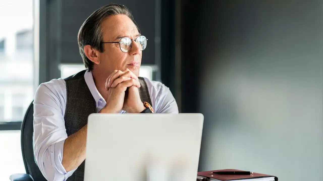 Older man in glasses and a vest, sitting at a desk with a laptop, looking away thoughtfully.