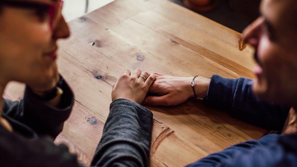 Close-up of a couple holding hands across a wooden table, seen from a high angle.
