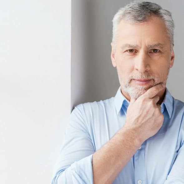 A middle-aged man with gray hair is posing by a window with his hand on his chin.